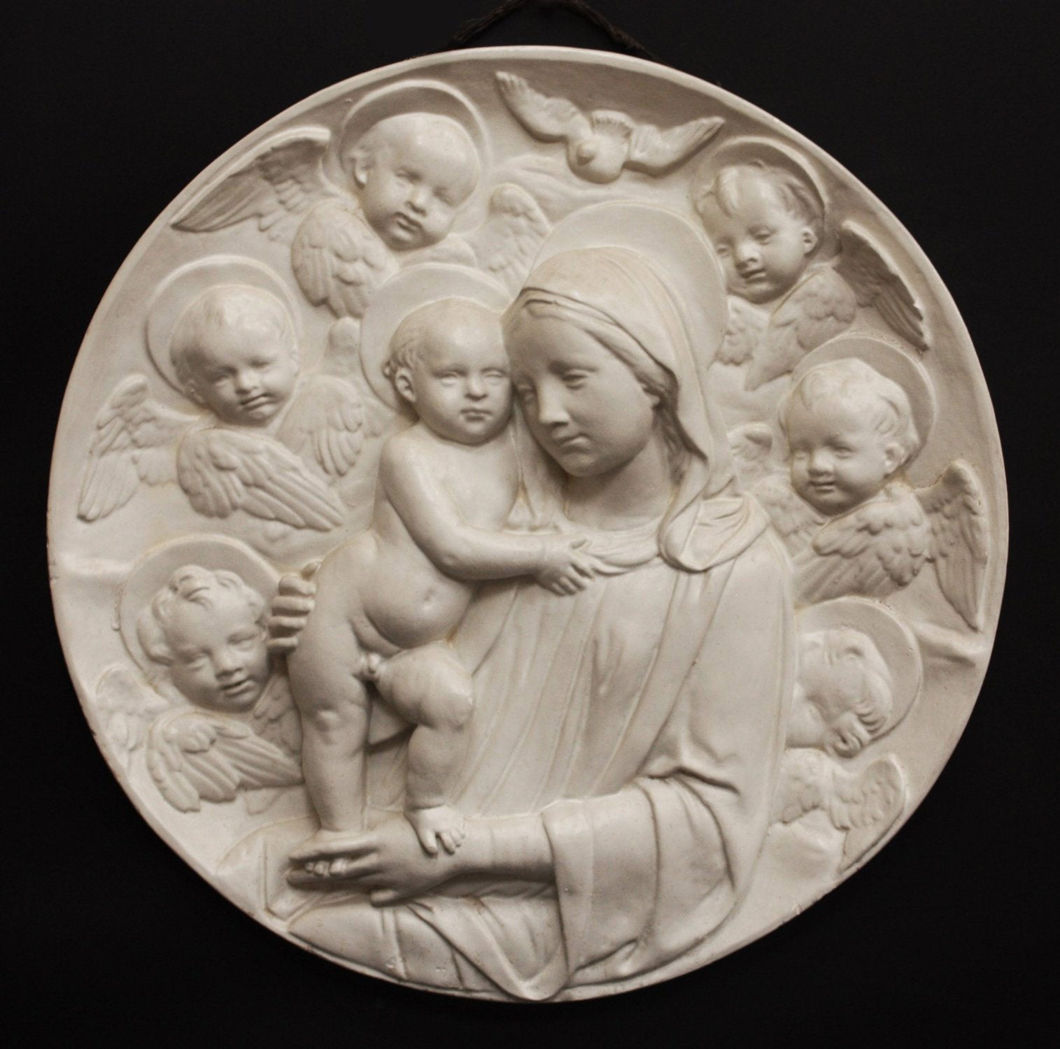 photo with black background of plaster cast relief of Madonna holding the Baby Jesus who&#39;s standing on her lap, and Angels encircling the round plaque