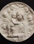 photo with black background of plaster cast relief of Madonna holding the Baby Jesus who's standing on her lap, and Angels encircling the round plaque