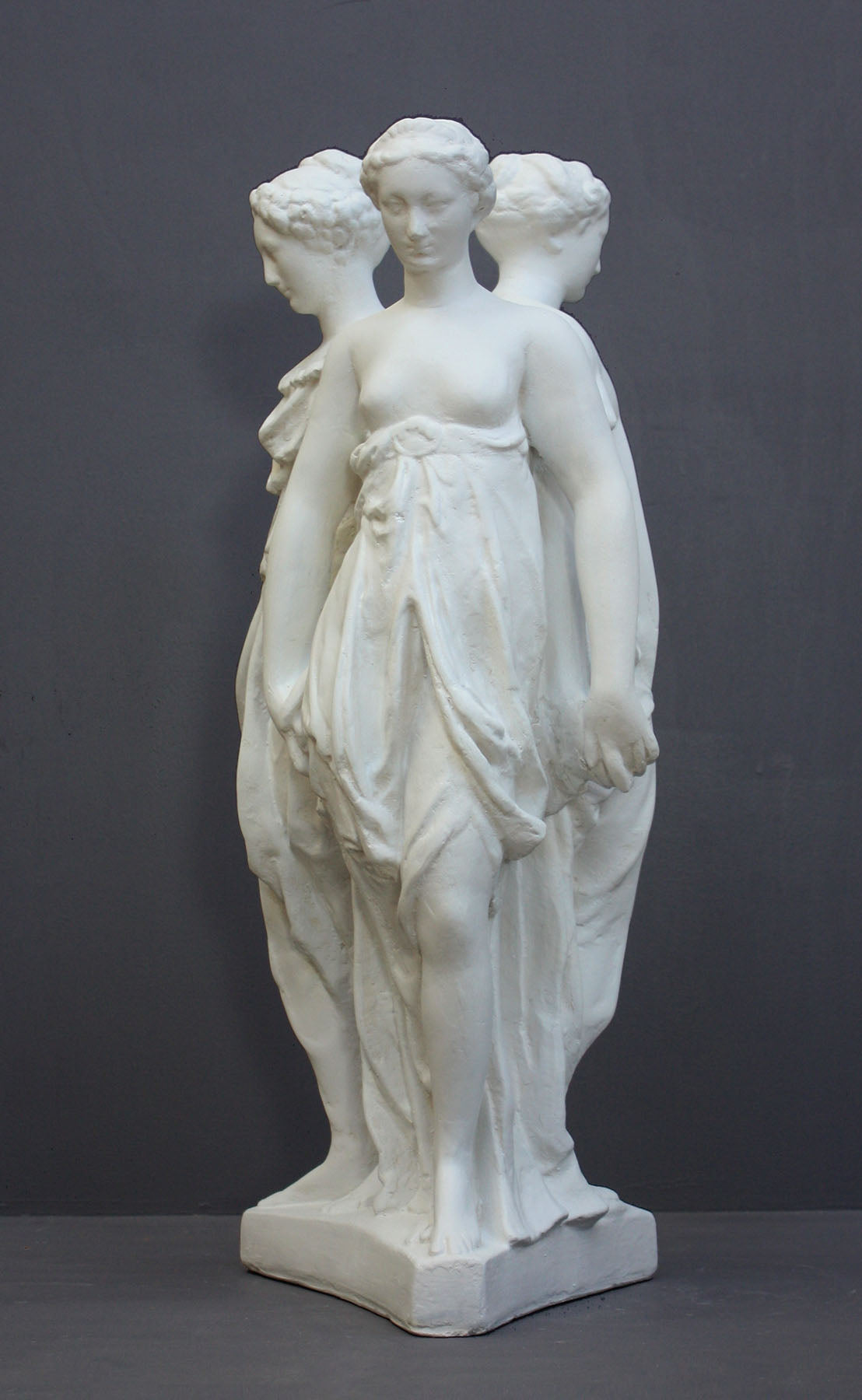 photo of white plaster cast sculpture of three robed females standing in a circle with their backs to each other against a gray background