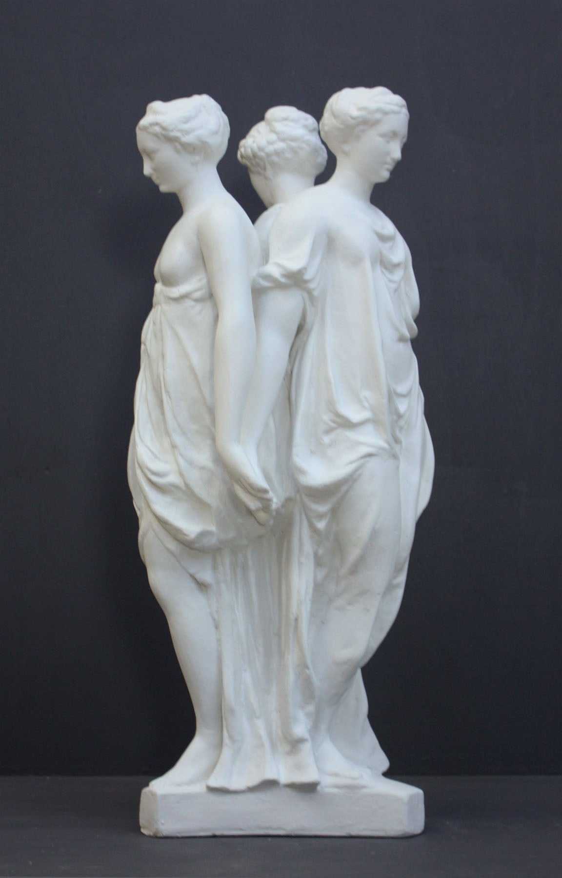 photo of white plaster cast sculpture of three robed females standing in a circle with their backs to each other against a gray background