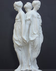 photo of white plaster cast sculpture of three robed females standing in a circle with their backs to each other against a gray background