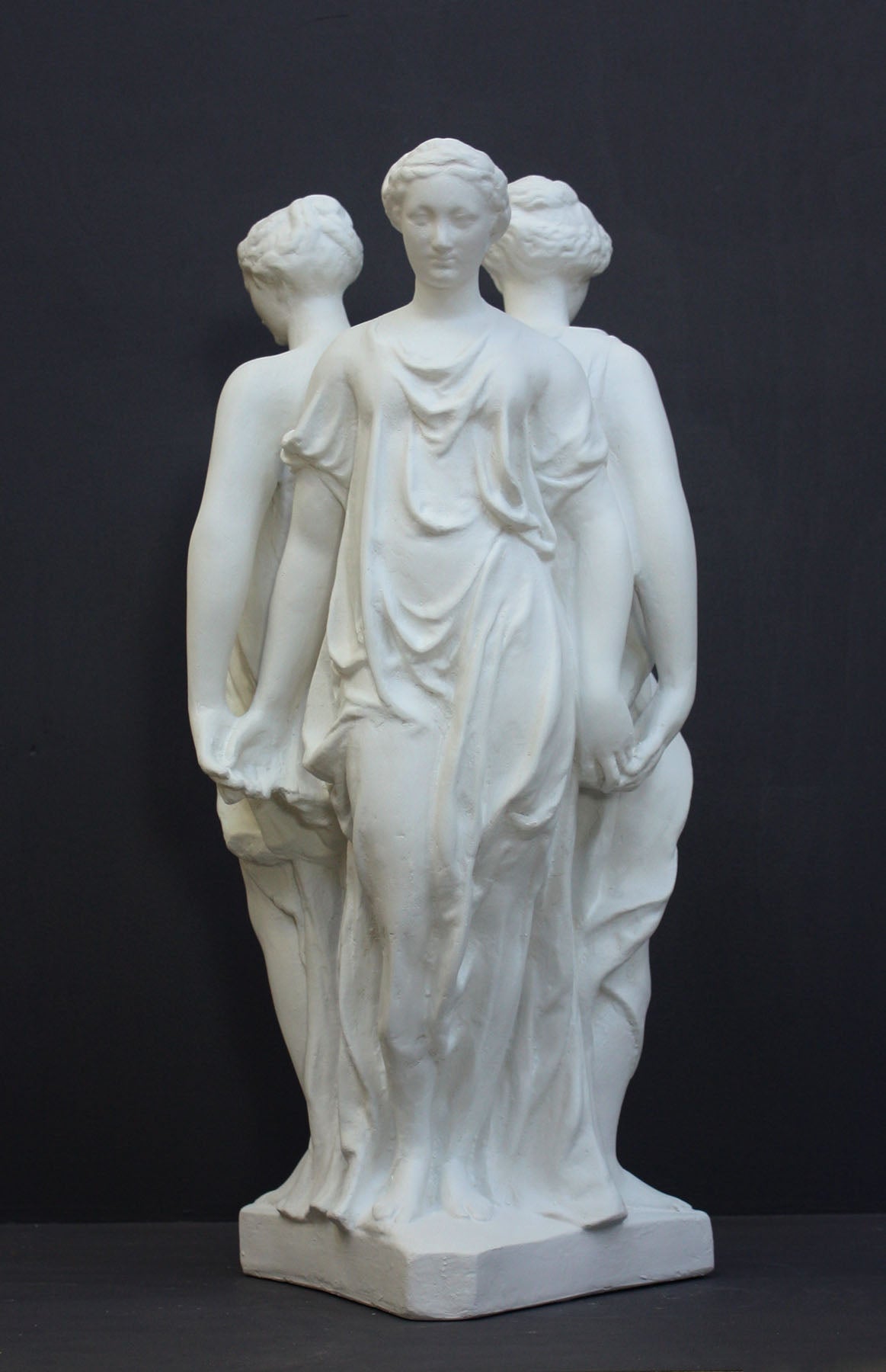 photo of white plaster cast sculpture of three robed females standing in a circle with their backs to each other against a gray background