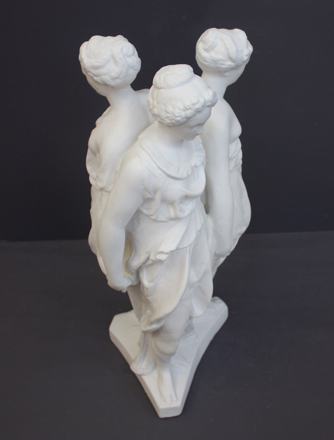 photo of white plaster cast sculpture of three robed females standing in a circle with their backs to each other against a gray background