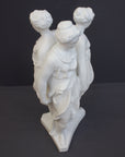 photo of white plaster cast sculpture of three robed females standing in a circle with their backs to each other against a gray background