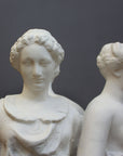 closeup photo of two female heads from white plaster cast sculpture of three robed females standing in a circle with their backs to each other against a gray background