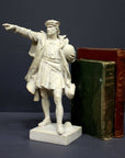 photo of plaster cast sculpture of standing man, namely Christopher Columbus, in robes and hat, pointing with right hand and holding a globe in his left arm with green and red books standing beside it against gray background