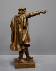 photo of bronze-colored plaster cast sculpture of standing man, namely Christopher Columbus, in robes and hat, pointing with right hand and holding a globe in his left arm against gray background