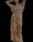 photo of plaster cast statue of a female in pink-orange tinted robes with her arms raised to her head against black background